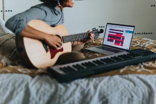 Sitting cross-legged on a bed, a person holds a guitar and appears to sing. Next to them is a musical keyboard and an open laptop.
