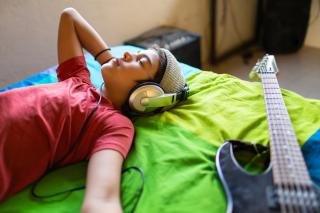 A young teen lies on their bed, eyes closed, listening to music on headphones. A guitar lies on the bed next to them.