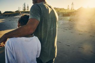 Seen from behind, at the beach, a father has his arm lovingly wrapped around his child's shoulder.