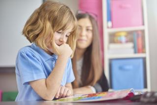 A child sits at a desk, reading, while in the background a teacher observes her.