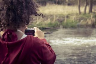 From behind, a child holds a camera at the edge of a river. In the distance, and through the camera screen, there's a heron faintly visible.