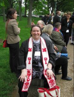 Rev. Cynthia Landrum, smiling on a park bench, wearing her ministerial stole. Clergy and other people crowd the walkway behind her.