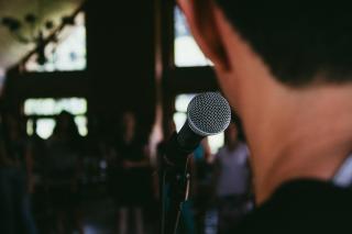 The back of a person's head as they speak into a microphone; in front of them, a blurred line of people