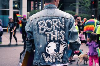 The back of a person's denim jacket says, "Born this way" in metal studs, with a unicorn. In the background, people wave rainbow flags.