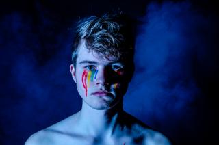 From the shoulders up, a white person gazes into the camera. Small stripes of paint, arranged in a rainbow pattern, run down their cheeks.