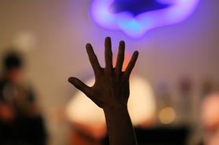 Over an out-of-focus background of musicians on a chancel, a single hand is raised in praise, fingers spread