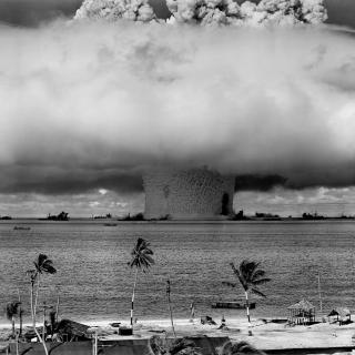 Grayscale photo of nuclear explosion's mushroom cloud on the horizon of a tropical sea, with a beach with palm trees and structures in the foreground