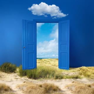 A blue double door is shown with some clouds above it and a sandy landscape in front. The doors are open to reveal a green field and cloudy blue sky beyond. 