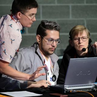 Three volunteers gather behind a laptop computer at a side table in General Session (the GA Tech Deck).