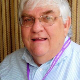 A portrait of Richard Nugent, an older white man. He is looking at the camera and has on glasses, a purple lanyard, and a blue striped shirt.