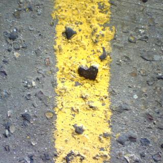 A close-up of a yellow stripe painted on rough asphalt. In the yellow stripe is embedded a heart-shaped stone.