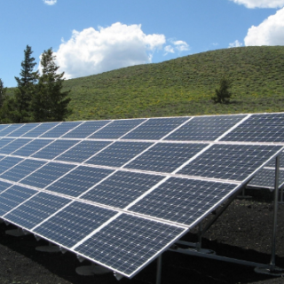 Photo of solar panels in front of a green hill with some trees and blue skies
