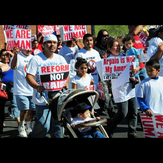 A man with children marches among others, many carrying signs like, 'We Are Human,' and 'Reform Not Raids.' Photo by George Pauk.