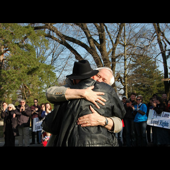 Two men embrace while rally attendees holding signs reading, 'Equal Love, Equal Rights,' look on and cheer. Photo courtesy Sue Ellen Tuttle.