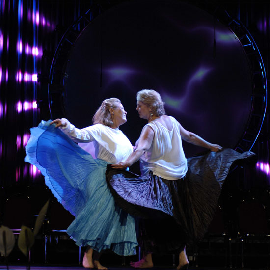 Two women in a stylized dance, their skirts flaring as they face each other in front of an elaborately lit stage. Photo by Nancy Pierce.