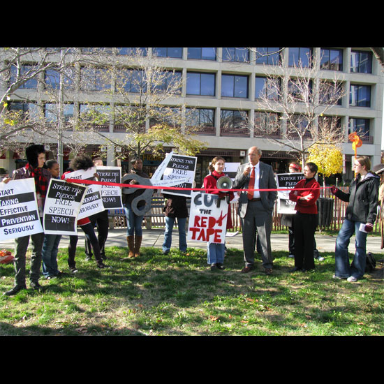 Sinkford speaking at the 2007 World AIDS Day event; people standing around a piece of red ribbon with signs reading, 'Cut the Red Tape.' Photo by Grace Garner/UUA.