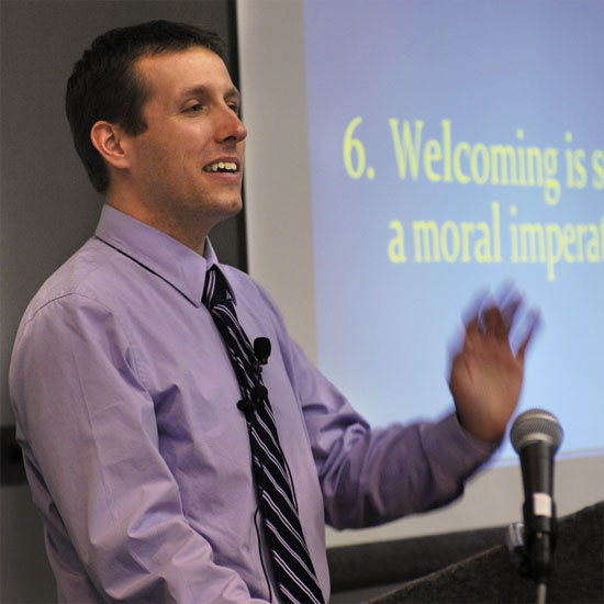 Thom Belote, speaking and gesturing in front of a presentation screen. Photo by Nancy Pierce.