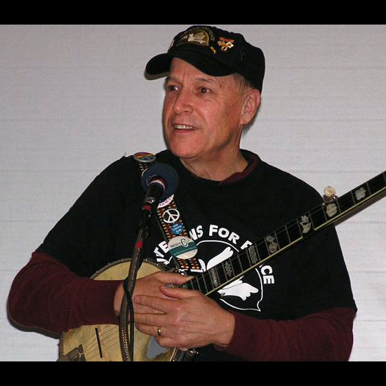 Pat Scanlon, wearing a shirt and a lot of buttons calling for peace, holds his banjo as he speaks to an audience. Photo by Art McDonald.