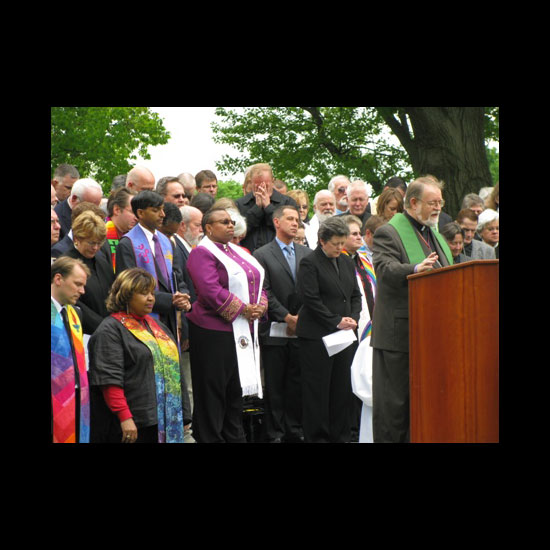A number of clergy bow their heads together in prayer behind a man speaking at a podium in an outdoor setting. Photo by Orelia Busch/UUA.