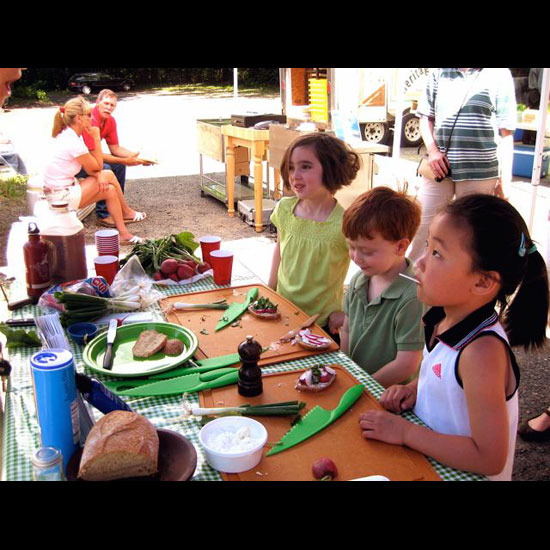 Children at a table with bread, vegetables, seasonings, cutting boards, and plastic knives. Photo by Barbara Harding.