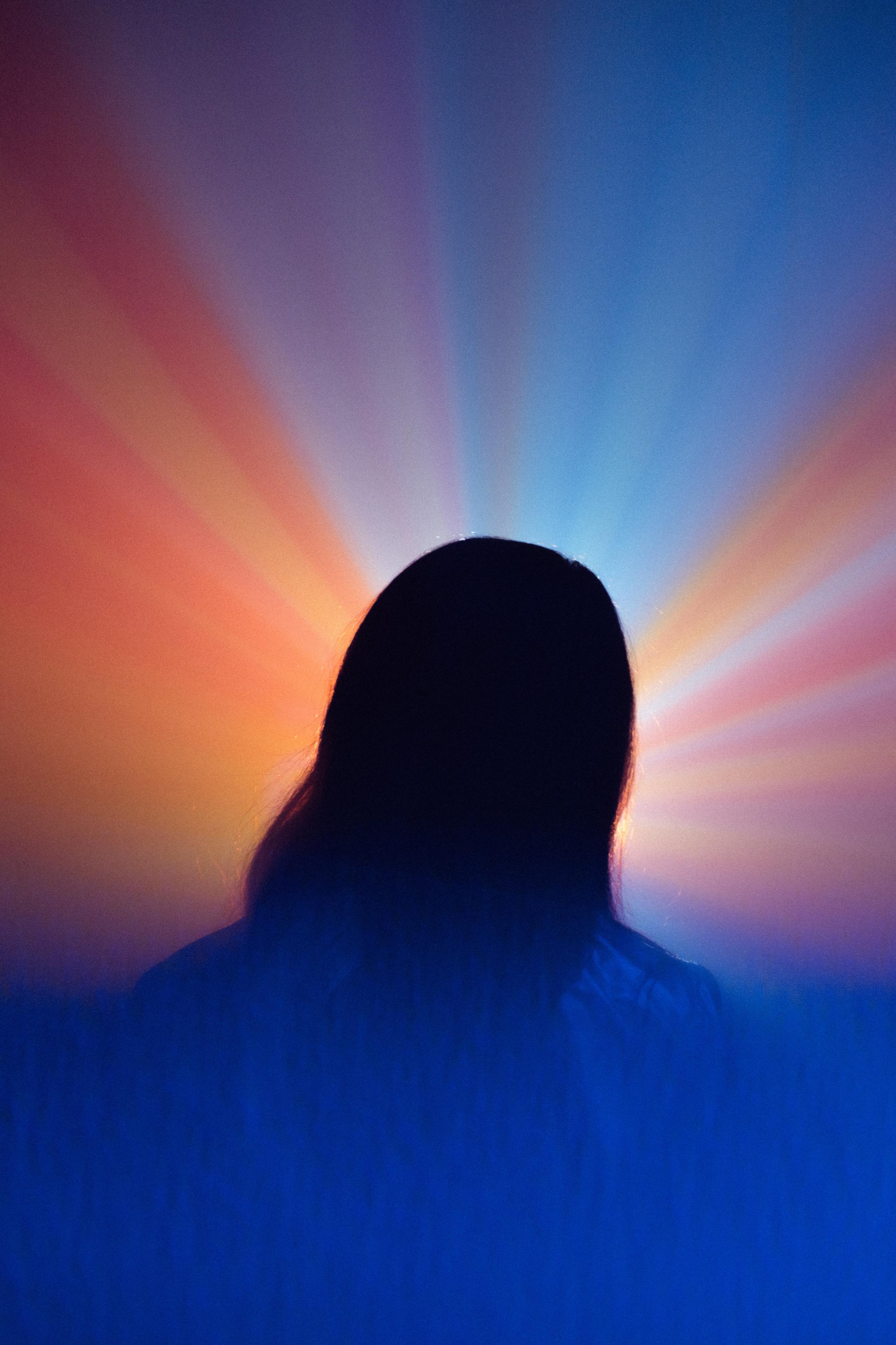 A person's head and shoulders, viewed from behind, with bright rays of colored light forming an aura.