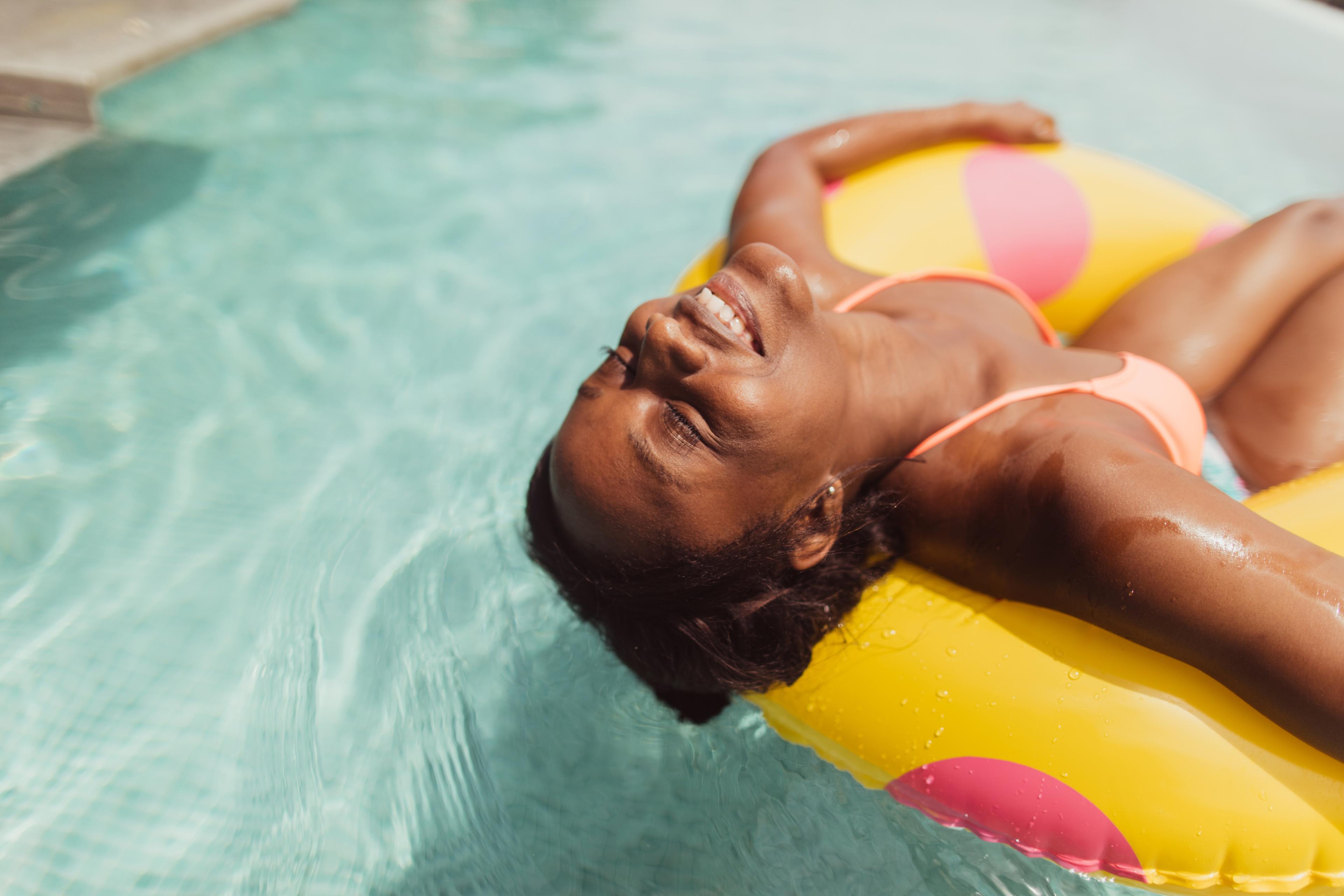A Black woman in a swimsuit smiles with closed eyes as she floats in a swimming pool on a bright yellow pool ring.