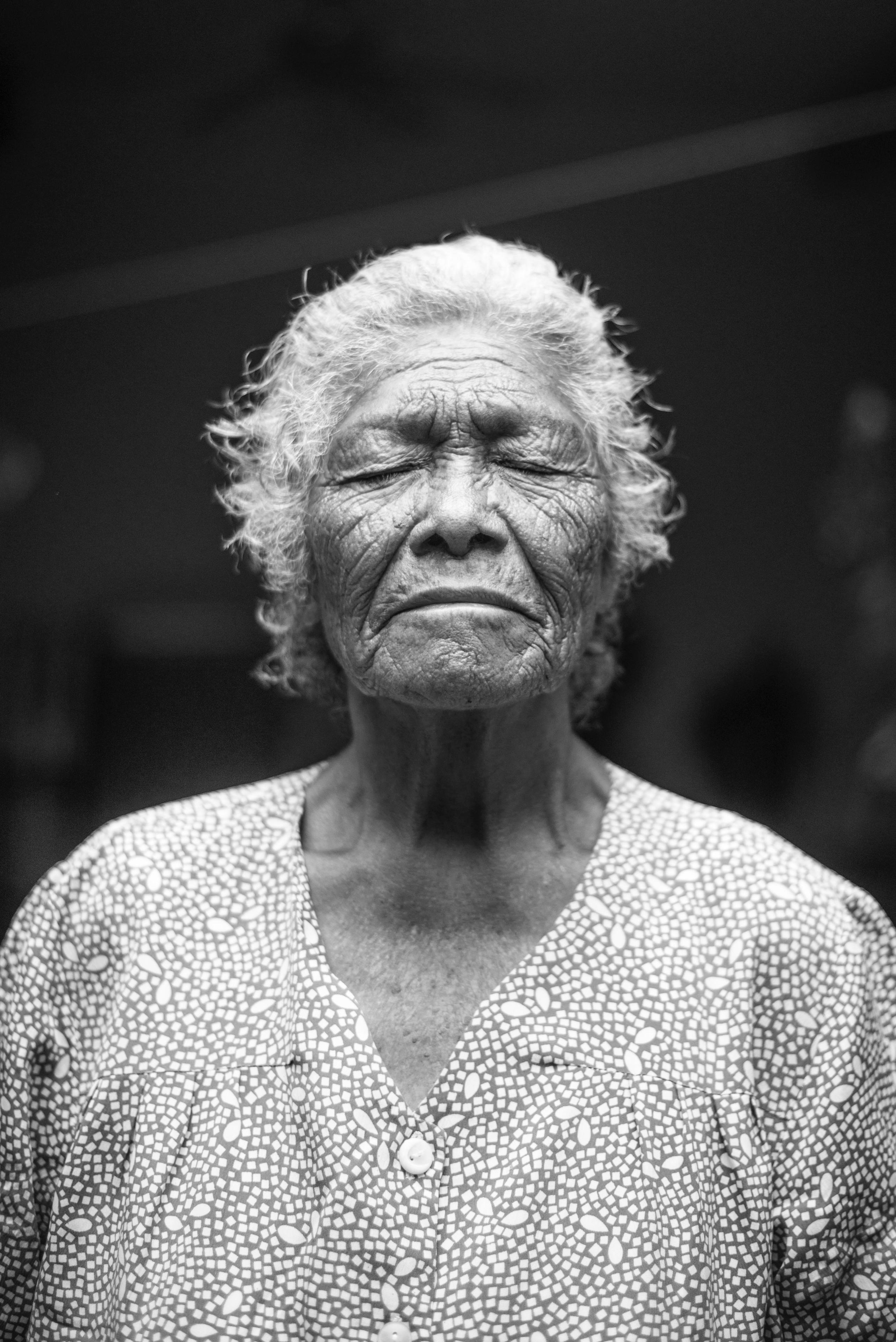 A black-and-white portrait of an older person with a deeply wrinkled face with closed eyes and white hair