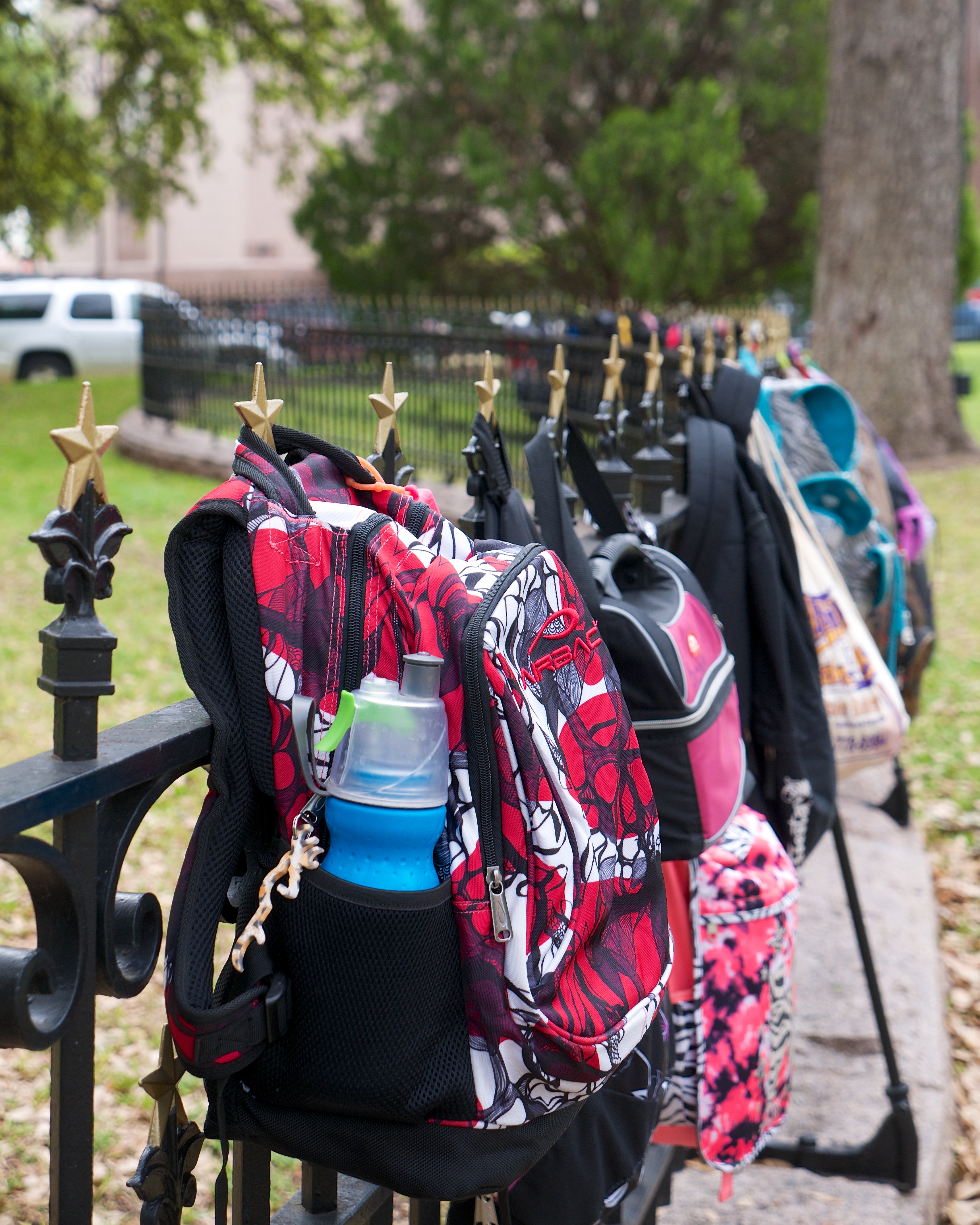 A range of schoolchildren's backpacks are hung on a wrought-iron fence