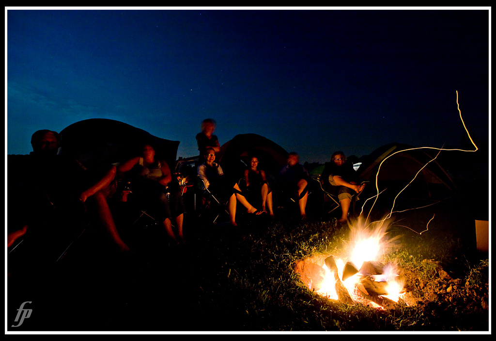 Against an indigo sky, people sit gazing into a sparking campfire.