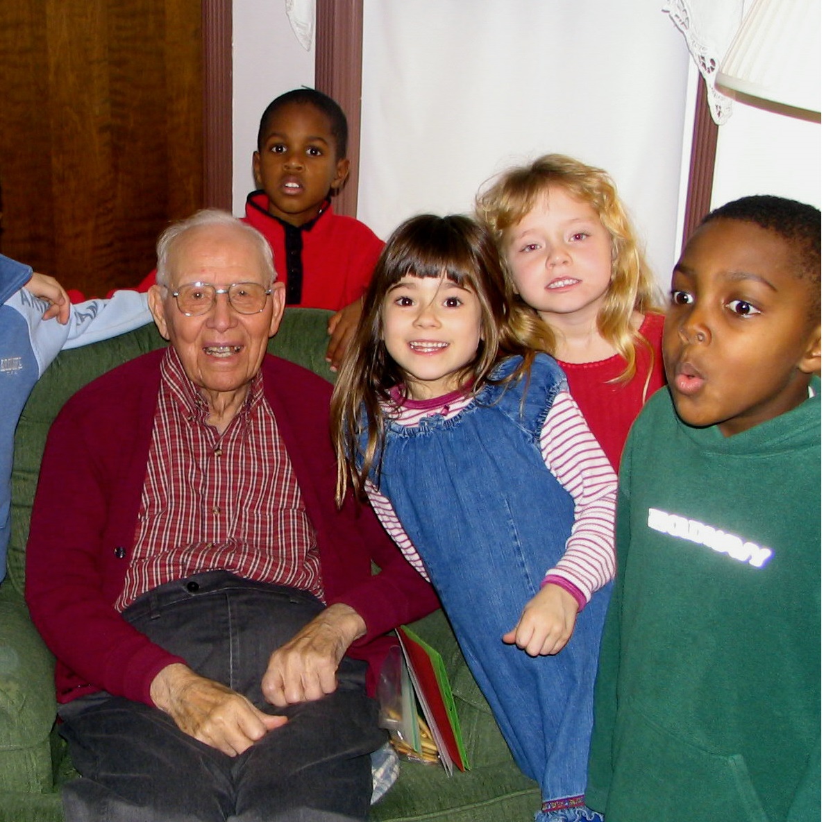An elderly white man sits in a cozy green chair, smiling, surrounded by children around seven years old, two white girls and two black boys part of a larger group of kids.