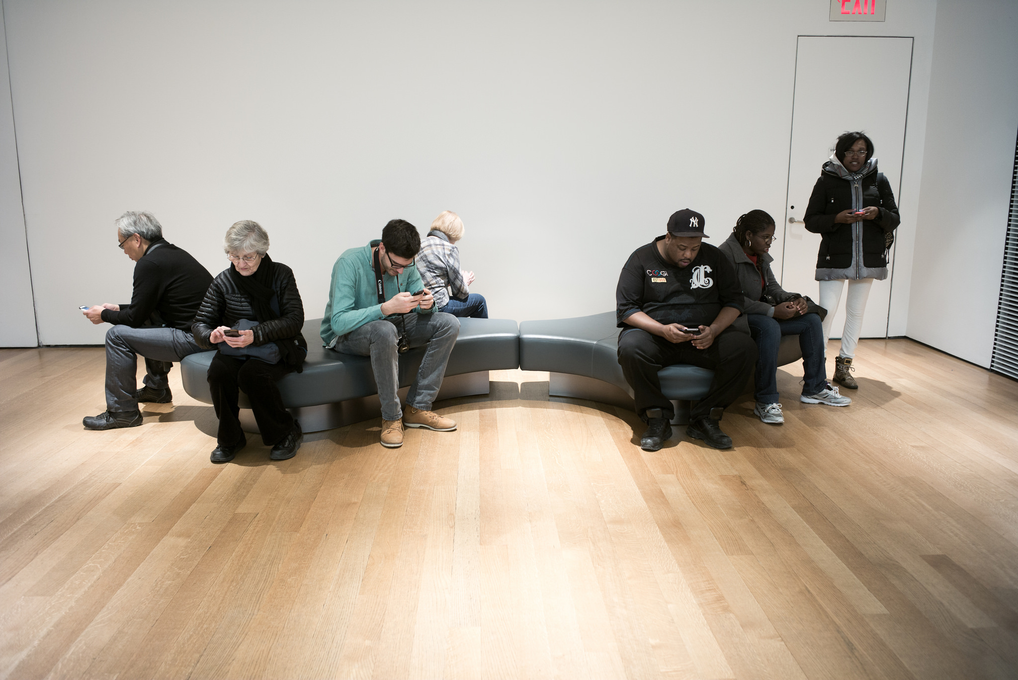 On and around a museum bench, seven different people each bows their head over their respective smartphone.