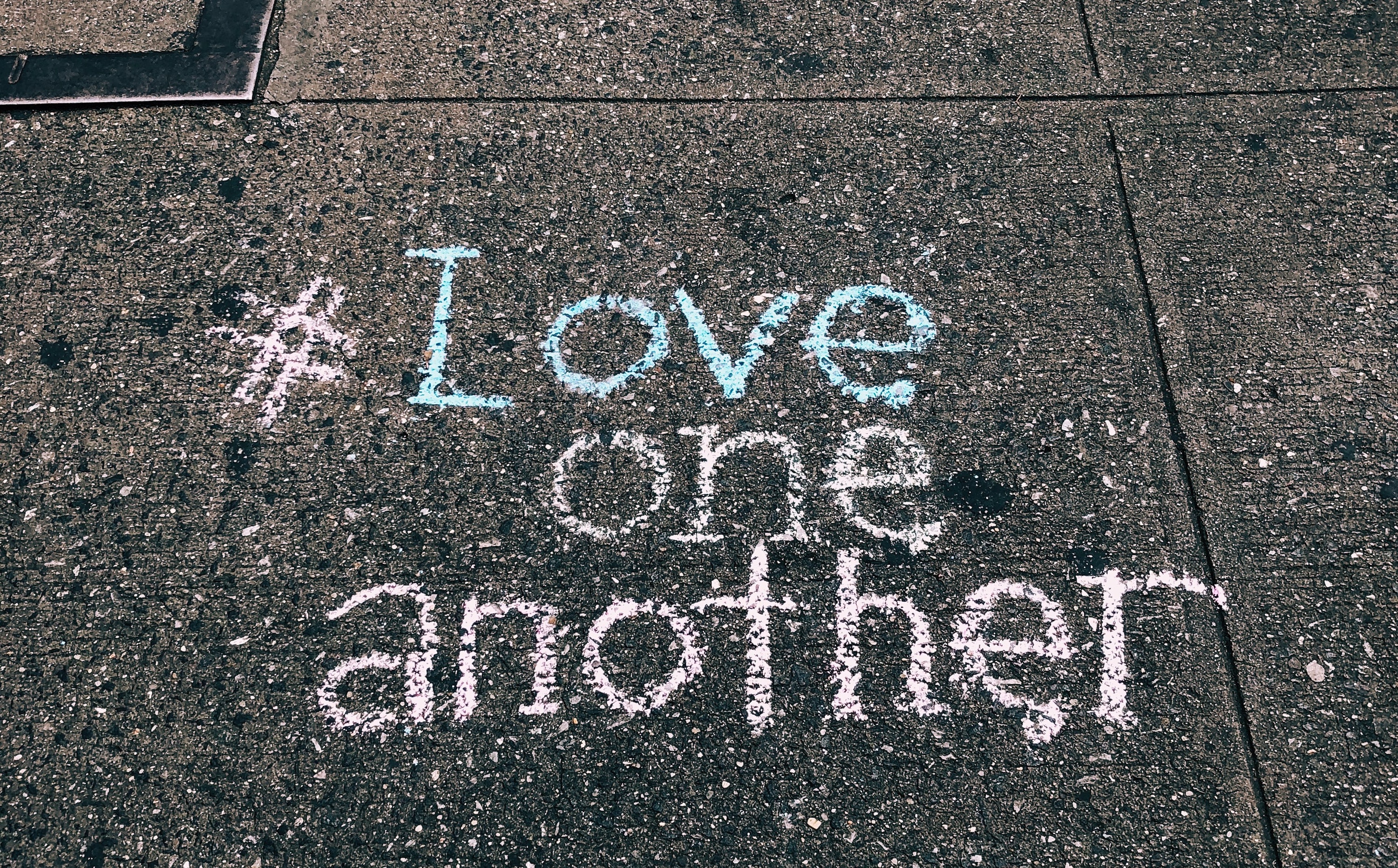 On pavement, the message written in pastel chalk, (hashtag) love one another. 