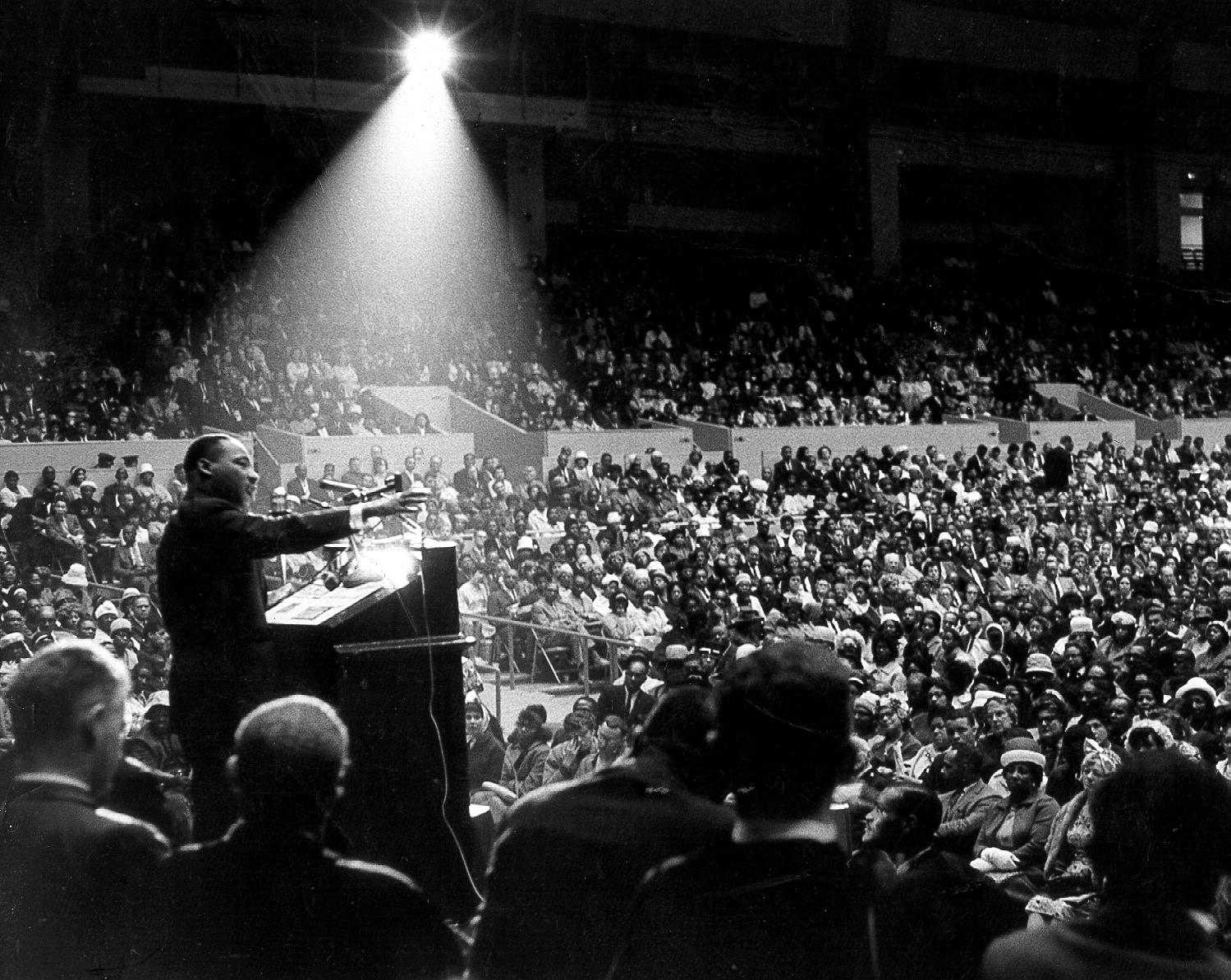 Martin Luther King, Jr. speaking to a crowd in San Francisco on June 30, 1964. In this black-and-white photograph, the humidity of the evening has cause a beam of light to appear surrounding King, as he gestures from a lectern.