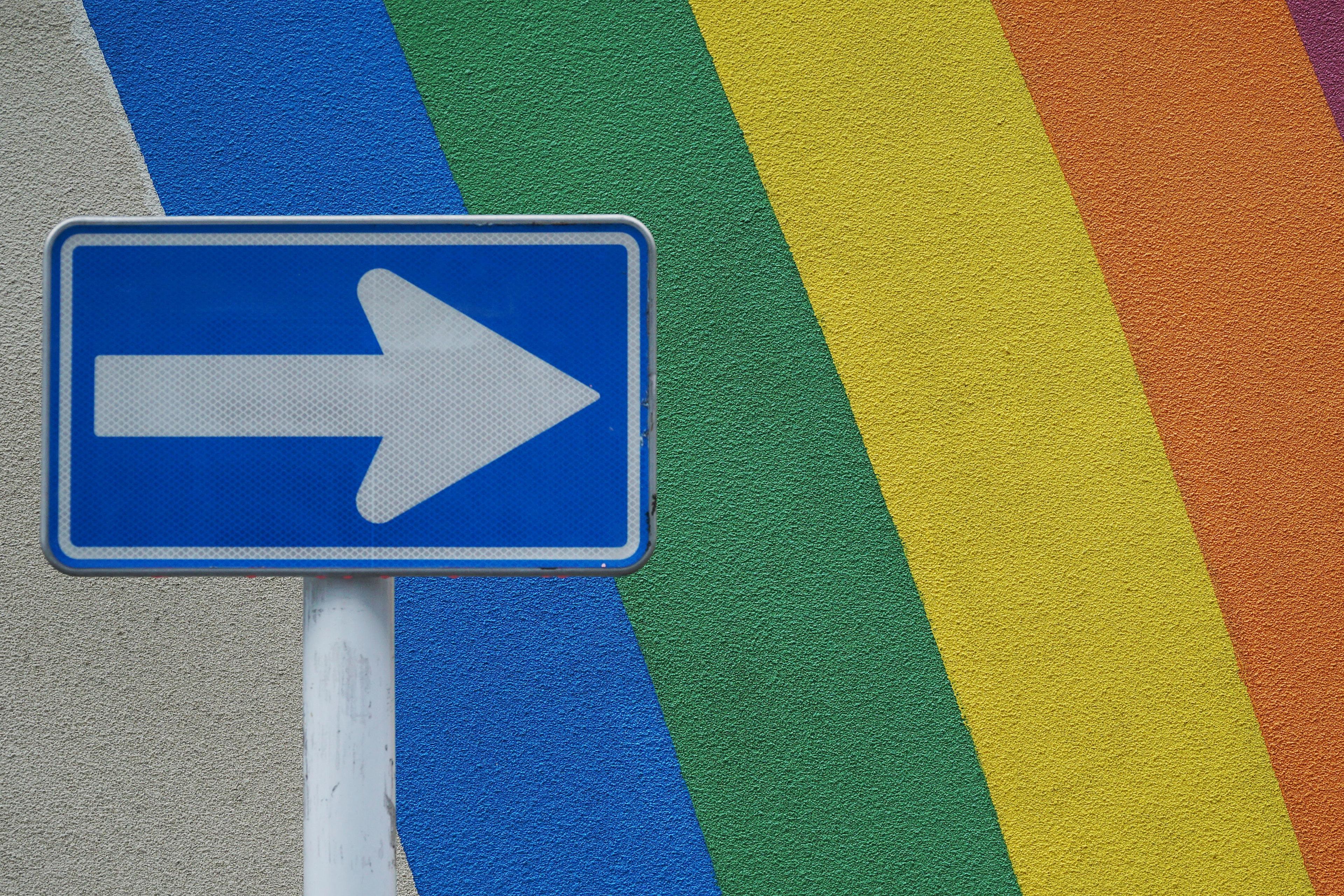 A blue arrow on a sign, against rainbow stripes painted on a wall