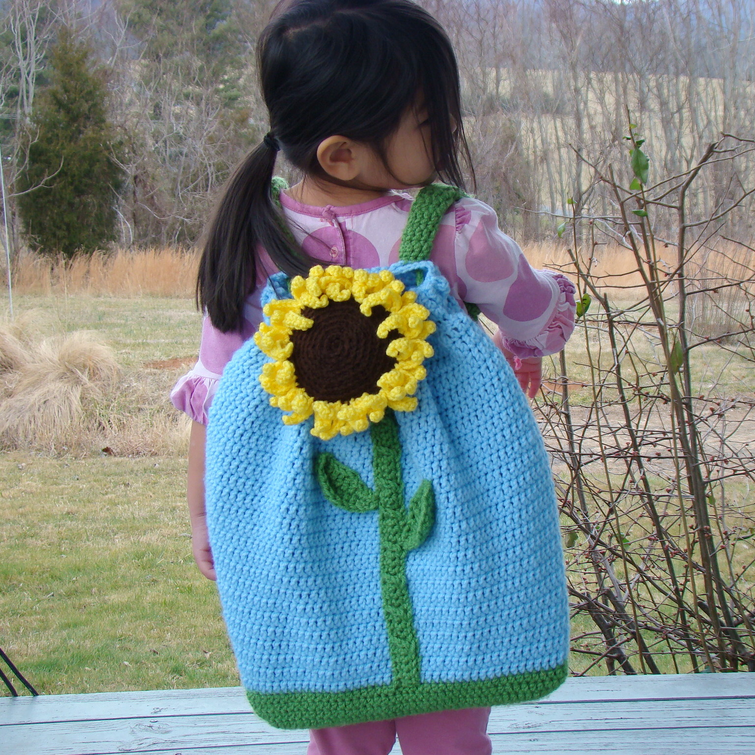 A small child, back to the camera, wearing a handmade crocheted blue backpack with a large sunflower on it.
