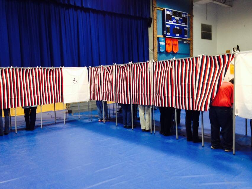 Inside of a school gymnasium, red-white-&-blue curtains hang along a string of voting booths, with some voters visible.