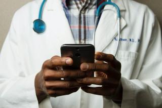 A person with dark skin, wearing a white doctor's coat and a stethoscope around their neck, holds a cell phone in their hands.