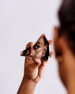 A black person holds a jagged piece of mirror, looking into it, as the camera captures the reflection oftheir eye.
