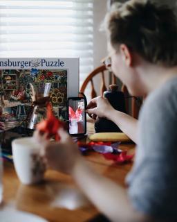 A person in a gray shirt is doing a jigsaw puzzle, holding up a piece to someone else on a video call on a cell phone.