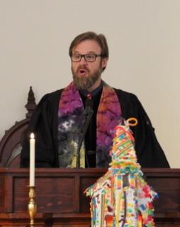 Rev. Paul, speaks from the pulpit, with a gingerbread house visible on the altar below.