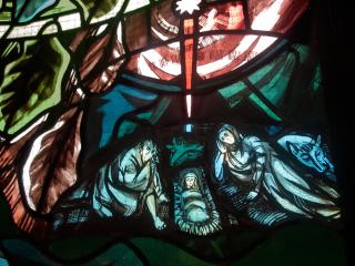 A close-up of a stained glass panel, in reds and greens, depicting Joseph and Mary reclining next to a manger holding Jesus