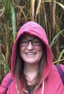 Laura Conkle smiles in an outdoor photo. They are wearing glasses and a pink hoodie.