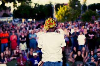 A Black woman, with her back to the camera, speaks into a microphone in front of a large audience outdoors, which appears to be a rally or protest.