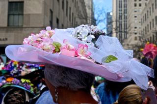 Outdoors, and seen from behind, a person with short gray hair wears a colorful hat with tulle and pink flowers.