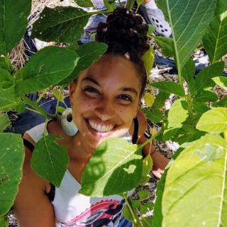 Photo of Leah Penniman smiling at the camera from behind green leaves