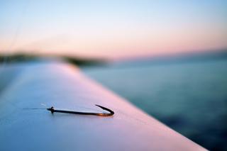 A sharp, gleaming fish hook lies on the edge of a boat, with a body of water visible in the background.