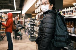 In a grocery store, a person wearing a mask looks off-camera while two other masked shoppers shop behind them