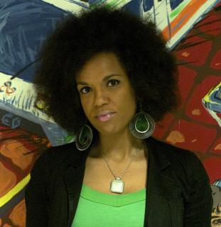 A black woman with afro and large green earrings in front of a colorful wall.