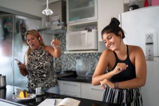 In a kitchen, a mother with an iPod and headphones dances and sings while her teenage daughter also dances.