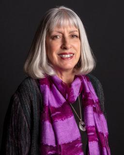 A photo of Lynn Sabourin, a white woman with silver, shoulder length hair, smiling and wearing a dark top and a purple stole